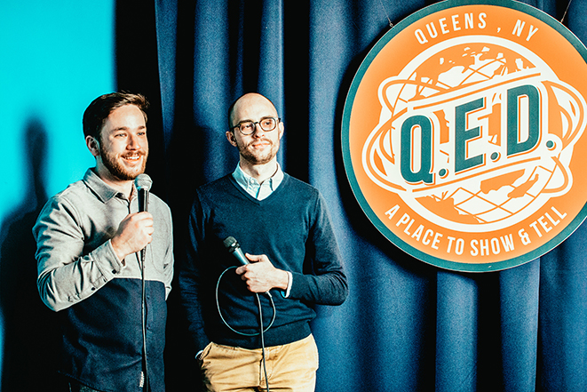 Patrick Cartelli and Evan Barden holding microphones on the QED stage.